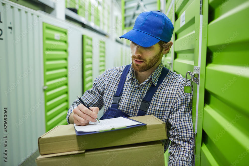 Young bearded worker in cap holding cardboard box and marking the parcel while working in storage room Young bearded worker in cap holding cardboard box and marking the parcel while working in storage room