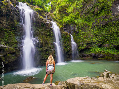 Blonde girl admires Three Bears Falls in Maui, Hawaii Hana Highway - Upper Waikani Falls. Road to Hana connects Kahului to the town of Hana Over 59 bridges, 620 curves, tropical rainforest.