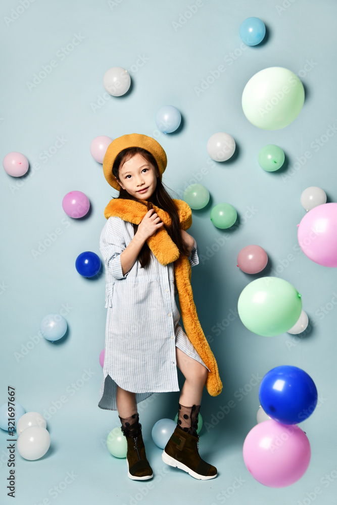 Fototapeta premium Little brunette asian girl in oversized shirt dress, brown beret and scarf, She posing with balloons on blue background.