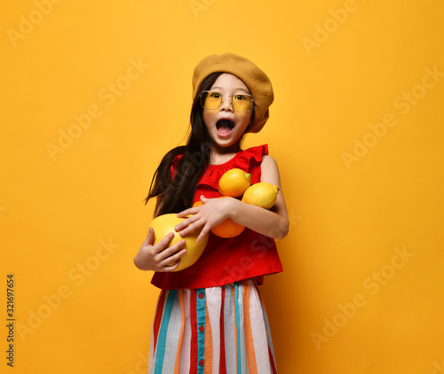 Little asian girl in sunglasses  brown beret  red blouse  colored skirt. Excited  holding pomelo  oranges and lemons. Studio shot.