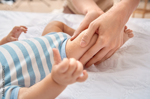Baby Care. Young mother doing pediatric massage massaging legs of little son lying on bed close-up