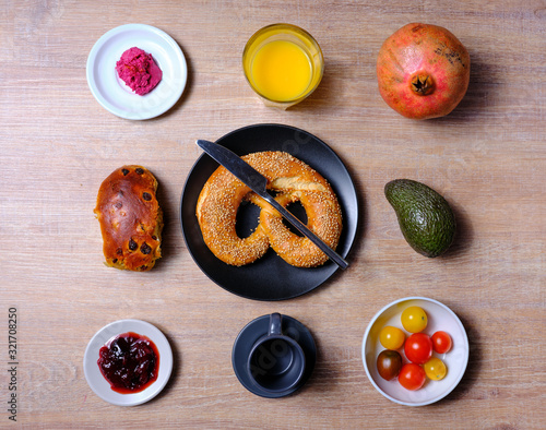 Pretzel, sweet tea-cake, marmelade, avocado, tomatoes, a pomegranate and a glass of orange juice, symetrically arranged on a wooden table on white and black tableware photo