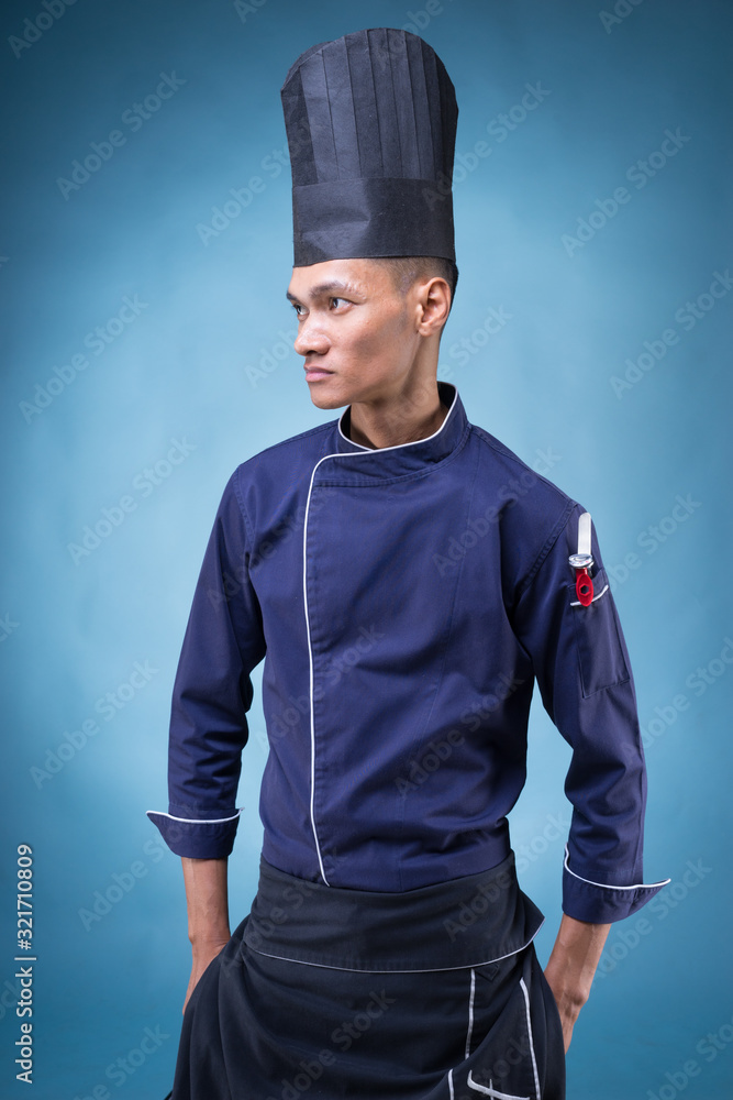 A portrait of an Asian executive chef in his blue uniform, black apron and black hat on blue background A portrait of an Asian executive chef in his blue uniform, black apron and black hat on blue background