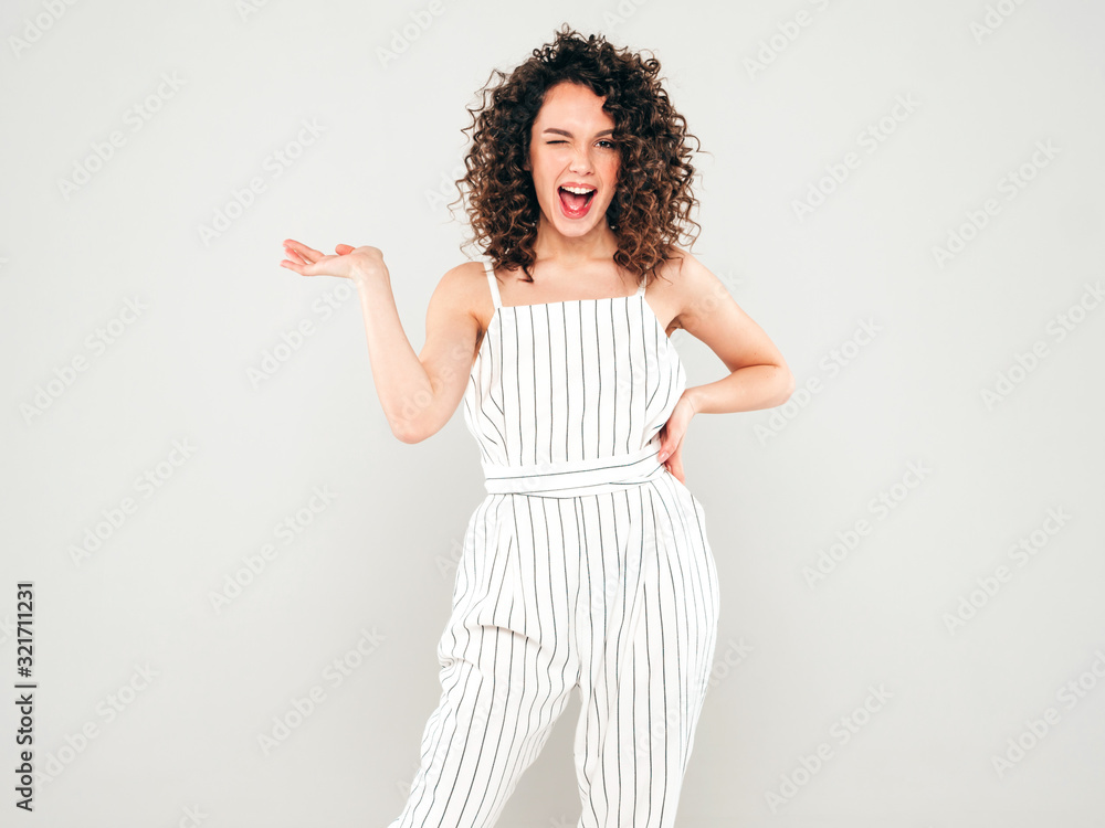 Portrait of beautiful smiling model with afro curls hairstyle dressed in summer hipster clothes.Sexy carefree girl posing in studio on gray background.Trendy woman shows something on her hand Portrait of beautiful smiling model with afro curls hairstyle dressed in summer hipster clothes.Sexy carefree girl posing in studio on gray background.Trendy woman shows something on her hand