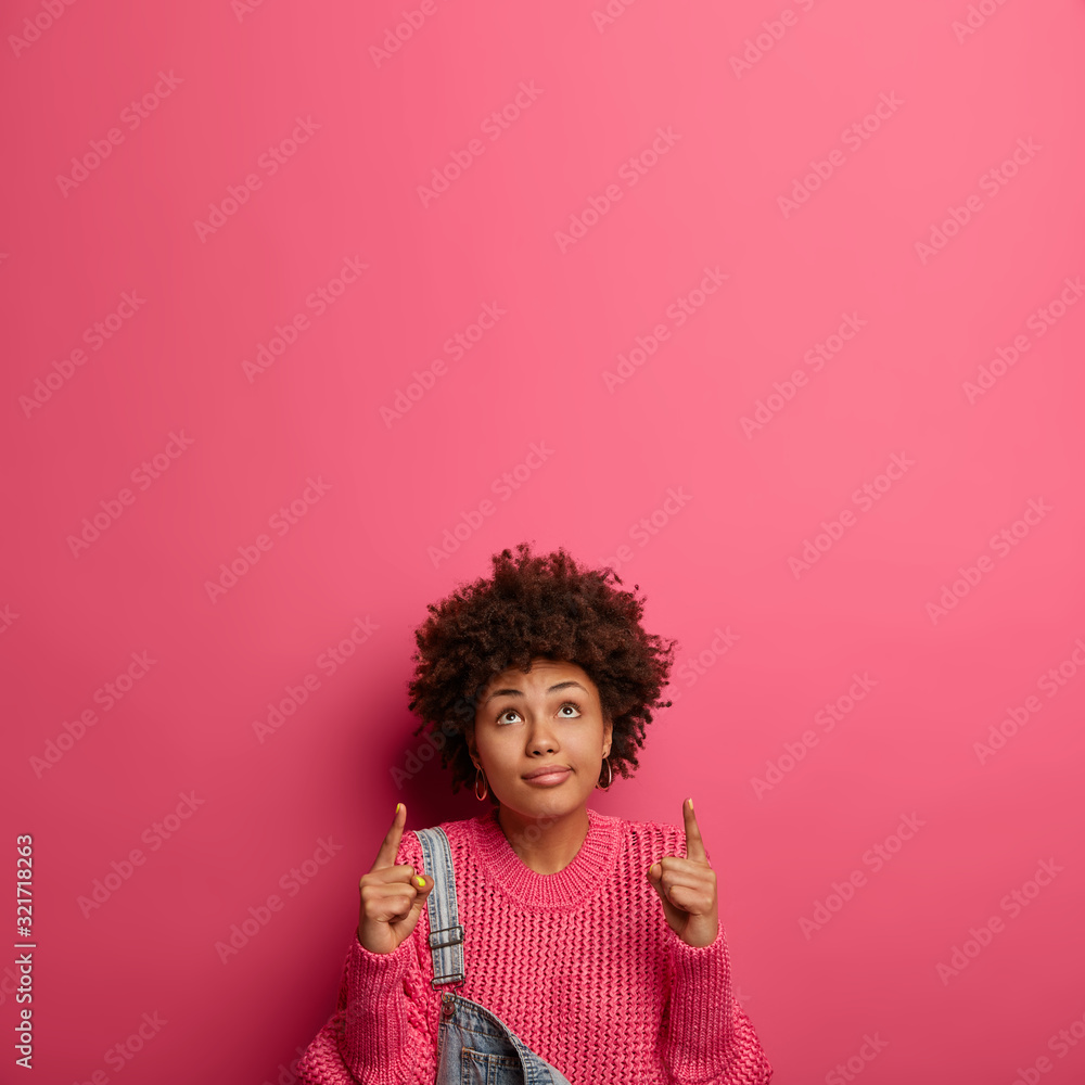 Curious dark skinned woman in modern outfit, looks and points up, checks out promotion, keeps gaze above, isolated over crimson background with empty space for your advertisement or information