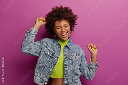 Triumphing happy curly woman celebrates finding new job, rasises arms in clenched fists, dressed in fashionable clothes, closes eyes with pleasure, enjoys loud music, dances carefree indoor. photo