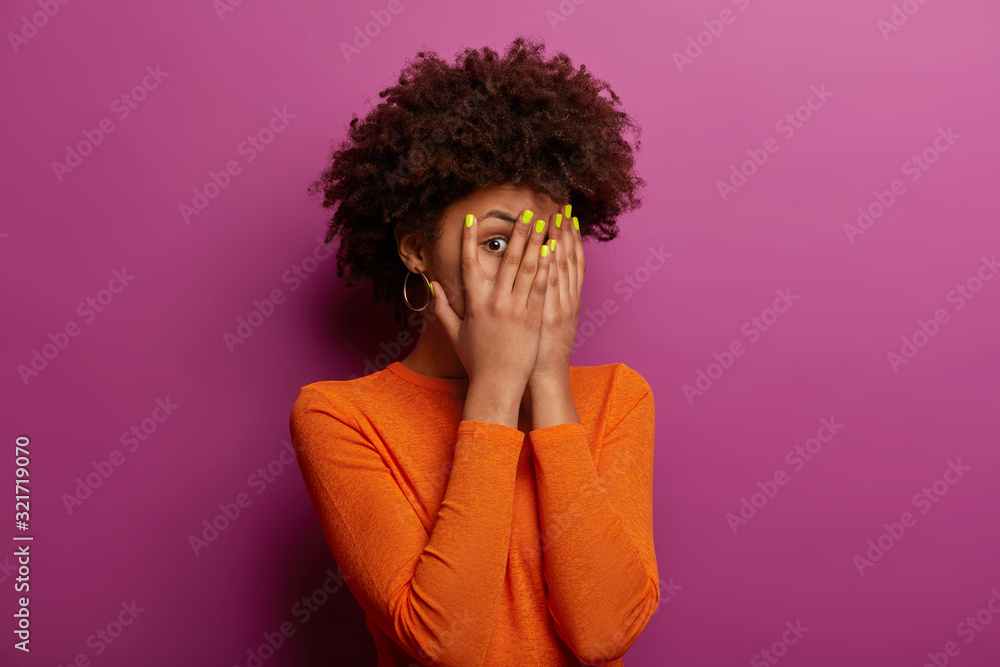 Young ethnic woman peeks through fingers, hides face with palms, has bright manicure, afraids of seeing something, wears casual orange sweater, stands against purple wall eager to see prepared present Young ethnic woman peeks through fingers, hides face with palms, has bright manicure, afraids of seeing something, wears casual orange sweater, stands against purple wall eager to see prepared present