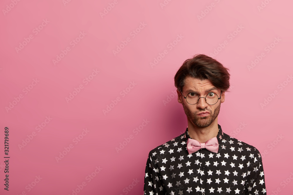 Photo of displeased unshaven man with trendy haircut, has gloomy face expression, wears round spectacles, stylish shirt with bowtie, stands against pink wall, free space left for your advert Photo of displeased unshaven man with trendy haircut, has gloomy face expression, wears round spectacles, stylish shirt with bowtie, stands against pink wall, free space left for your advert