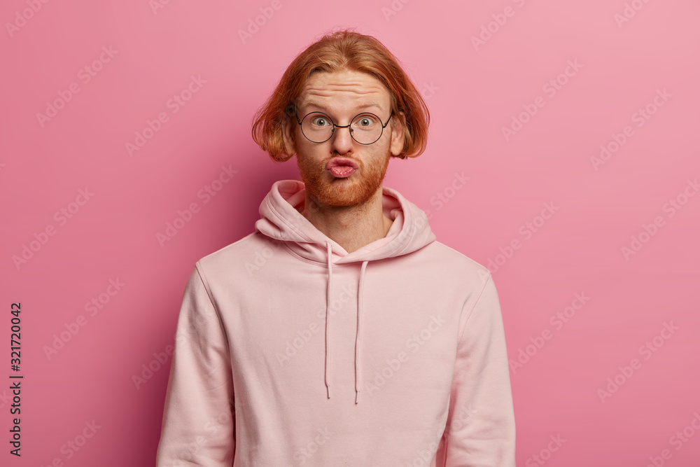 Portrait of funny bearded man with bob hairstyle, pouts lips and makes grimace, looks surprisingly through optical glasses, wears sweatshirt, poses against pink background. Human face expressions Portrait of funny bearded man with bob hairstyle, pouts lips and makes grimace, looks surprisingly through optical glasses, wears sweatshirt, poses against pink background. Human face expressions