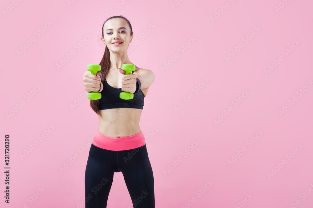 A young athletic woman, with a smile, in black sports top, doing aerobics on pink background with green light dumbbells. A young athletic woman, with a smile, in black sports top, doing aerobics on pink background with green light dumbbells.