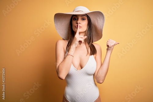 Young beautiful brunette woman on vacation wearing swimsuit and summer hat asking to be quiet with finger on lips pointing with hand to the side. Silence and secret concept.