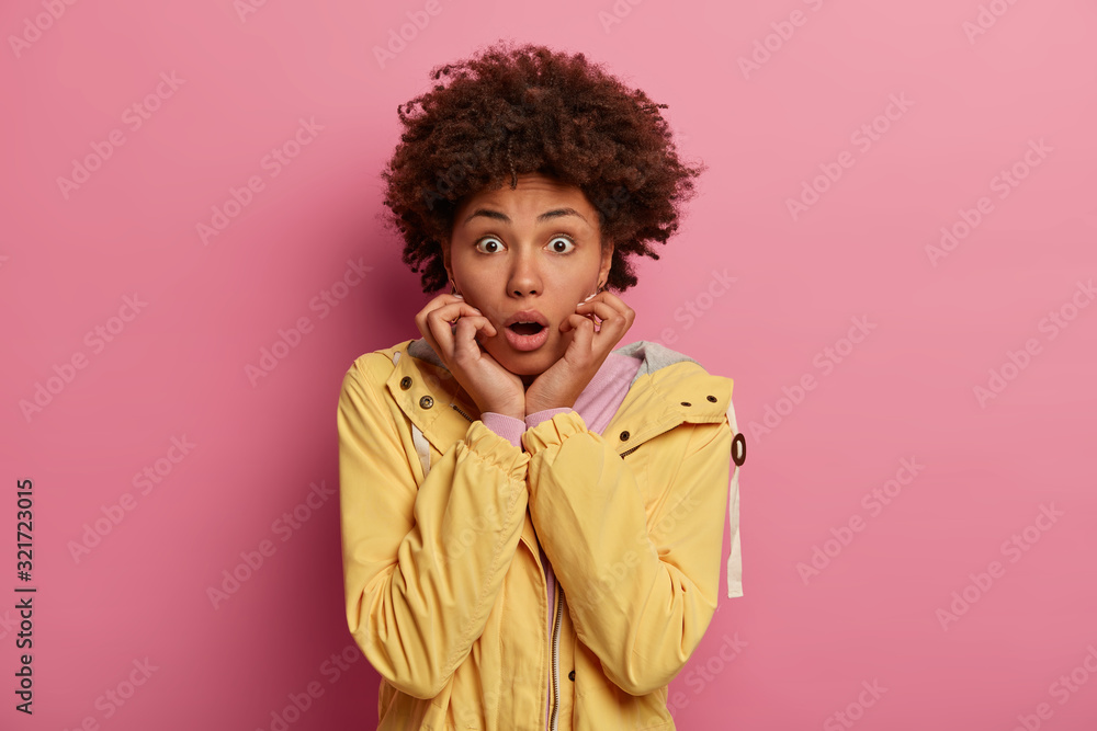 Horizontal shot of shocked amazed Afro American woman keeps mouth opened, gasps with fear, notices something strange and very scary, wears yellow windbreaker, isolated over rosy studio background Horizontal shot of shocked amazed Afro American woman keeps mouth opened, gasps with fear, notices something strange and very scary, wears yellow windbreaker, isolated over rosy studio background