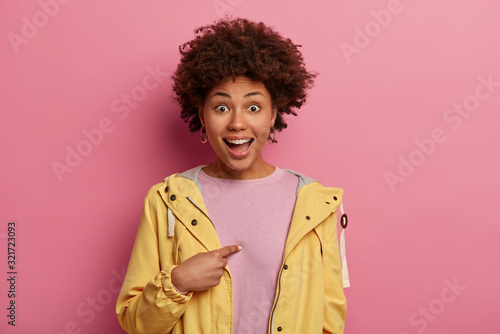 Really me? Surprised curly haird woman points at herself, cannot believe she is chosen, wears yellow anorak, has positive mood, isolated on pink background, opens mouth and eyes with surprisement