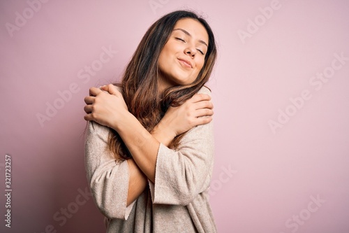 Young beautiful brunette woman wearing casual sweater standing over pink background Hugging oneself happy and positive, smiling confident. Self love and self care photo
