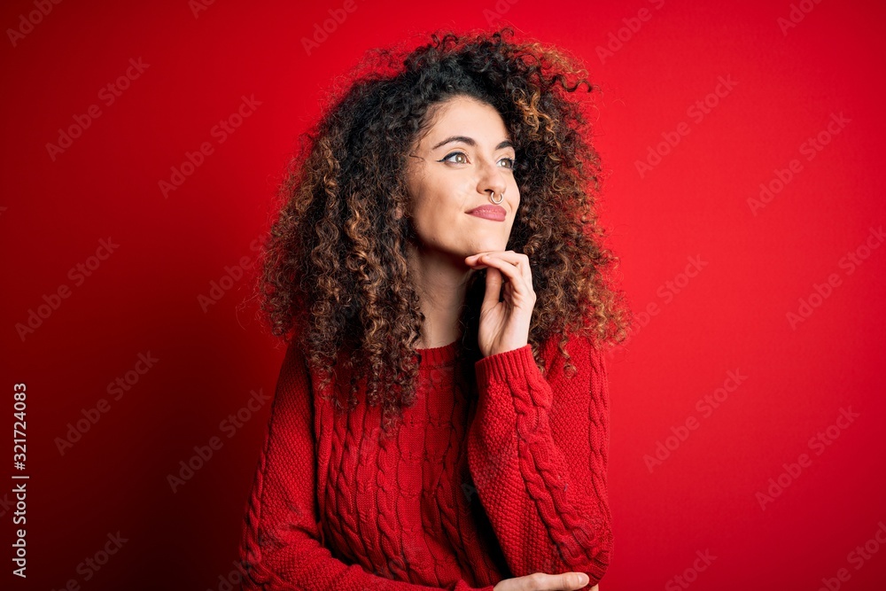 Young beautiful woman with curly hair and piercing wearing casual red sweater with hand on chin thinking about question, pensive expression. Smiling and thoughtful face. Doubt concept. Young beautiful woman with curly hair and piercing wearing casual red sweater with hand on chin thinking about question, pensive expression. Smiling and thoughtful face. Doubt concept.