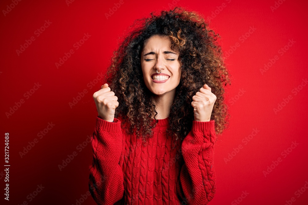 Young beautiful woman with curly hair and piercing wearing casual red sweater excited for success with arms raised and eyes closed celebrating victory smiling. Winner concept. Young beautiful woman with curly hair and piercing wearing casual red sweater excited for success with arms raised and eyes closed celebrating victory smiling. Winner concept.