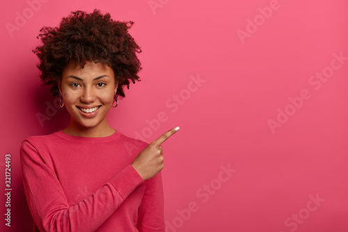 Pleasant looking cheerful Afro American woman points at upper right corner, discusses advertisement, gives advice what to buy, smiles gently, wears pink jumper. Find best price and discount there