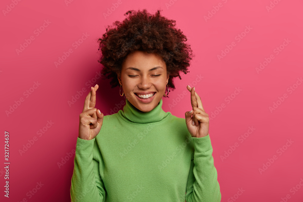 Hopeful happy woman smiles positively, makes wish and confident to win, crosses fingers for good luck, eagerly wishing success, closes eyes, wears green turtleneck, isolated over vibrant pink wall Hopeful happy woman smiles positively, makes wish and confident to win, crosses fingers for good luck, eagerly wishing success, closes eyes, wears green turtleneck, isolated over vibrant pink wall