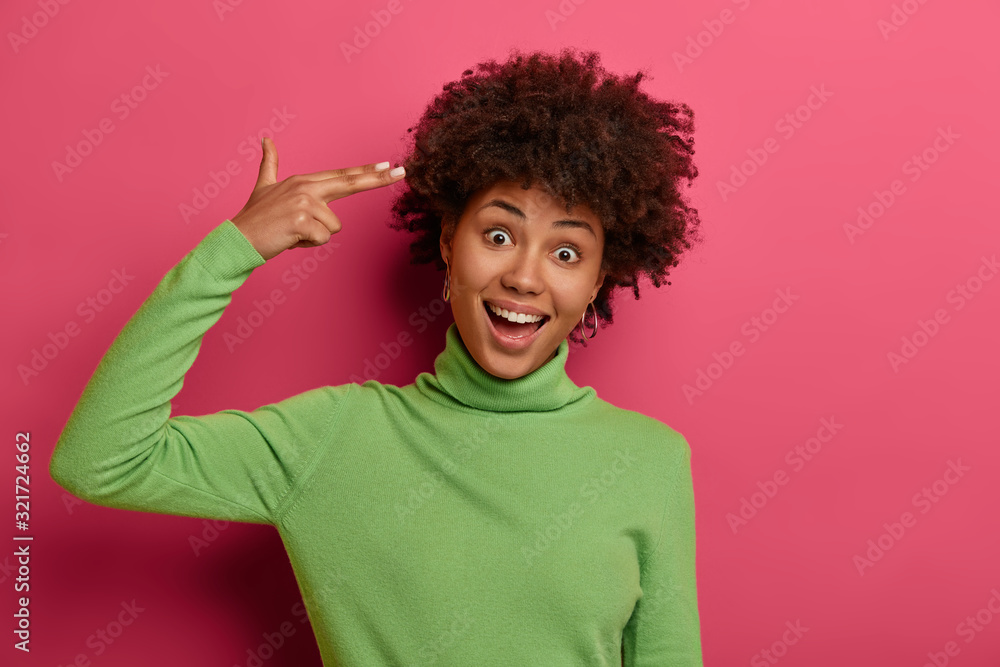 Portrait of joyous dark skinned woman shoots in temple with finger gun, tilts head and has funny expression, overjoyed look, natural curly hair, wears green poloneck, poses against crimson background. Portrait of joyous dark skinned woman shoots in temple with finger gun, tilts head and has funny expression, overjoyed look, natural curly hair, wears green poloneck, poses against crimson background.