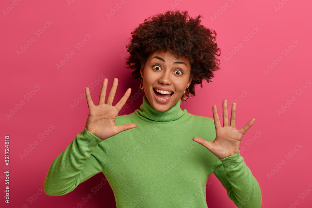 Isolated shot of positive Afro American woman keeps palms raised, smiles broadly with funny expression, wears green turtleneck, models against pink vivid background. People and emotions concept Isolated shot of positive Afro American woman keeps palms raised, smiles broadly with funny expression, wears green turtleneck, models against pink vivid background. People and emotions concept