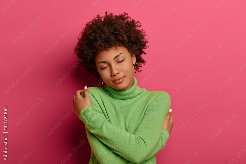 Tender romantic woman cuddles and holds herself in hugs, keeps eyes closed, likes her new green turtleneck, feels comfort, enjoys calm atmosphere, isolated on pink background. Just myself and I - obrazy, fototapety, plakaty 