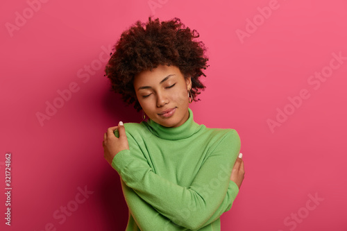Tender romantic woman cuddles and holds herself in hugs, keeps eyes closed, likes her new green turtleneck, feels comfort, enjoys calm atmosphere, isolated on pink background. Just myself and I