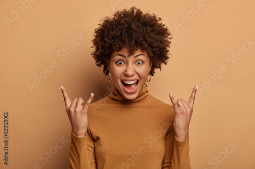 Rock n roll lives forever. Emotional dark skinned woman makes heavy metal sign, exclaims happily, attends cool concert or awesome party, enjoys favourite music, wears brown clothes, stands indoor.