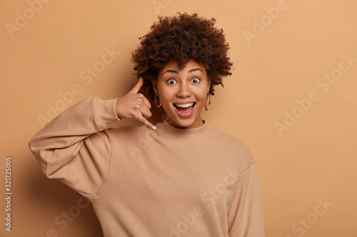 Positive beautiful woman asks to give her call sometime, makes phone gesture near ear, gives telephone number, wears casual beige sweatshirt, poses indoor. Body language ad emotions concept.