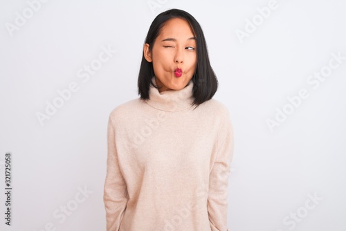 Young chinese woman wearing turtleneck sweater standing over isolated white background making fish face with lips, crazy and comical gesture. Funny expression. © Krakenimages.com