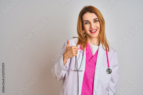 Redhead caucasian doctor woman wearing pink stethoscope over isolated background doing happy thumbs up gesture with hand. Approving expression looking at the camera showing success.