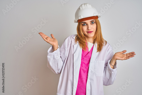 Redhead caucasian woman engineer wearing safety helmet over isolated background clueless and confused expression with arms and hands raised. Doubt concept.
