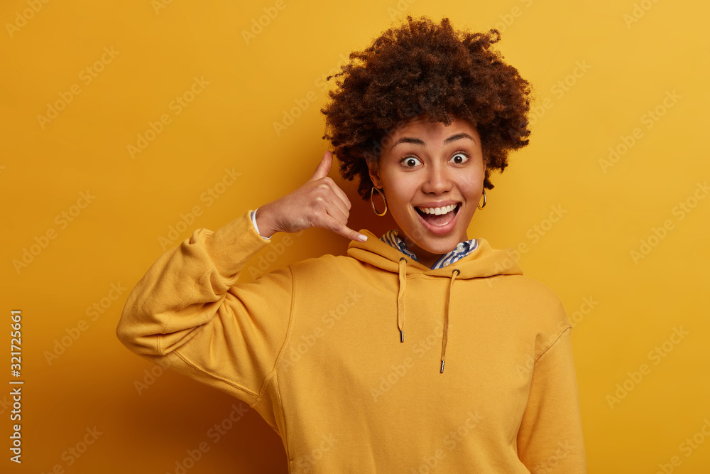 Funny glad Afro American female does phone gesture, keeps fingers like talking on smartphone, smiles broadly, wears sweatshirt, poses over vivid yellow wall. Communnicating concept. Call me back