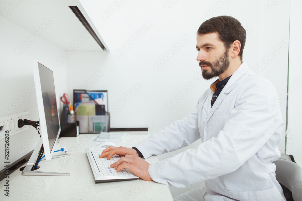Bearded mature dentist working on computer at his office. Male doctor typing on keyboard of a computer, working at the hospital Bearded mature dentist working on computer at his office. Male doctor typing on keyboard of a computer, working at the hospital