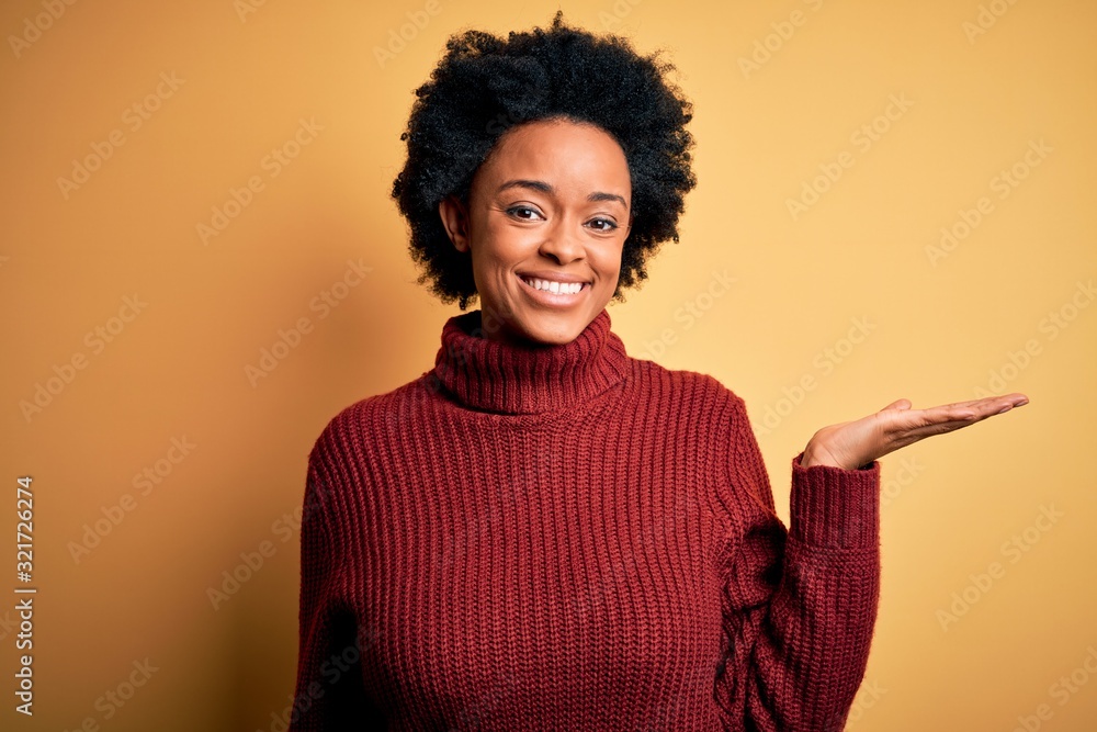 Young beautiful African American afro woman with curly hair wearing casual turtleneck sweater smiling cheerful presenting and pointing with palm of hand looking at the camera. Young beautiful African American afro woman with curly hair wearing casual turtleneck sweater smiling cheerful presenting and pointing with palm of hand looking at the camera.