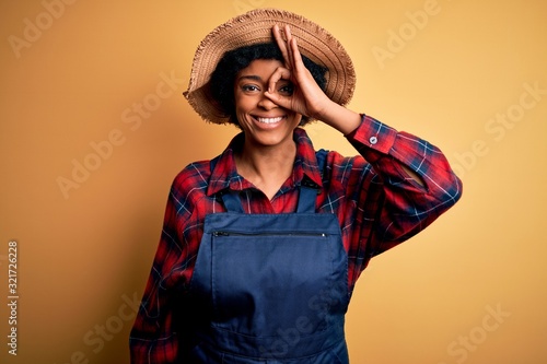 Young African American afro farmer woman with curly hair wearing apron and hat doing ok gesture with hand smiling, eye looking through fingers with happy face.