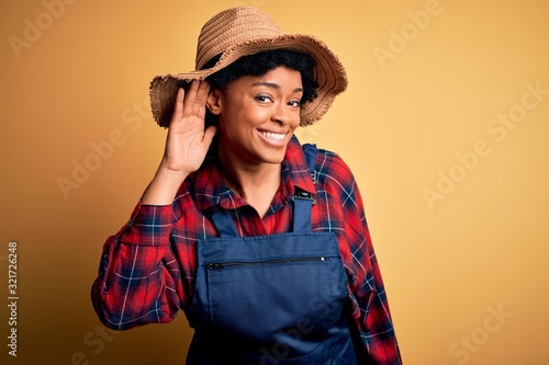 Young African American afro farmer woman with curly hair wearing apron and hat smiling with hand over ear listening an hearing to rumor or gossip. Deafness concept.