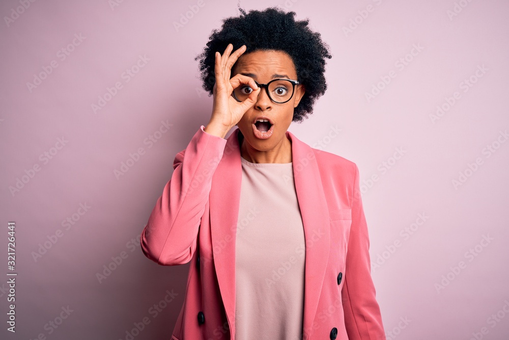 Young beautiful African American afro businesswoman with curly hair wearing pink jacket doing ok gesture shocked with surprised face, eye looking through fingers. Unbelieving expression. Young beautiful African American afro businesswoman with curly hair wearing pink jacket doing ok gesture shocked with surprised face, eye looking through fingers. Unbelieving expression.