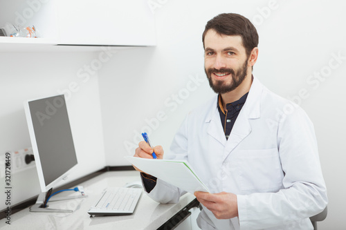 Happy mature male doctor smiling to the camera, sitting at his office. CHeerful practitioner doing paperwork at his clinic