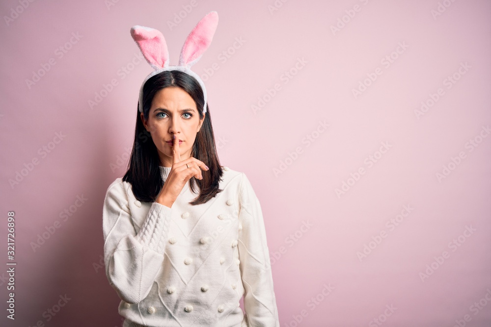 Young caucasian woman wearing cute easter rabbit ears over pink isolated background asking to be quiet with finger on lips. Silence and secret concept.
