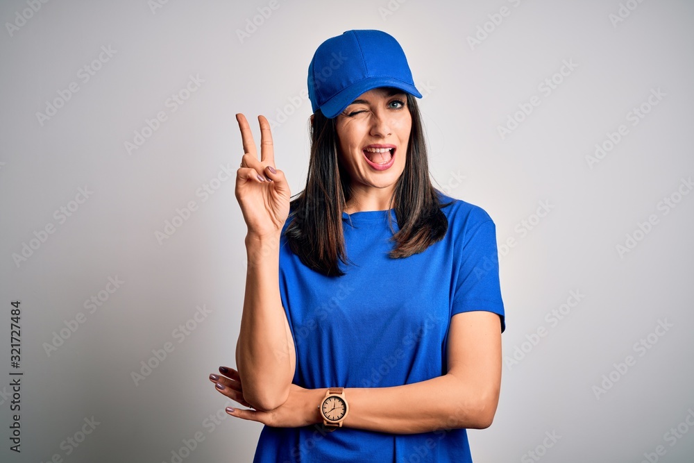 Young delivery woman with blue eyes wearing cap standing over blue background smiling with happy face winking at the camera doing victory sign. Number two. Young delivery woman with blue eyes wearing cap standing over blue background smiling with happy face winking at the camera doing victory sign. Number two.
