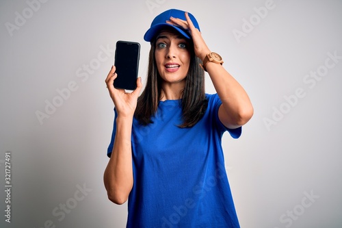 Young delivery woman with blue eyes wearing cap holding smartphone stressed with hand on head, shocked with shame and surprise face, angry and frustrated. Fear and upset for mistake.