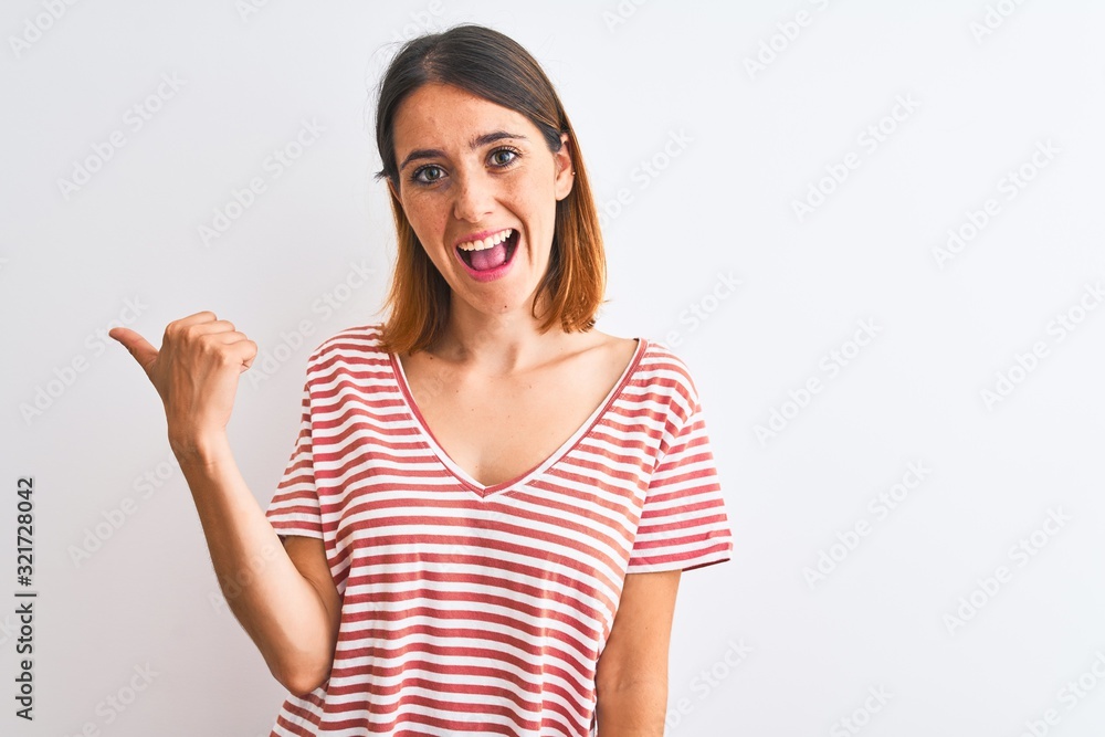 Beautiful redhead woman wearing casual striped red t-shirt over isolated background smiling with happy face looking and pointing to the side with thumb up. Beautiful redhead woman wearing casual striped red t-shirt over isolated background smiling with happy face looking and pointing to the side with thumb up.