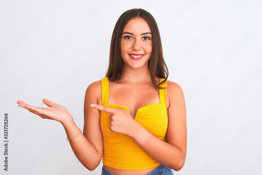 Young beautiful girl wearing yellow casual t-shirt standing over isolated white background amazed and smiling to the camera while presenting with hand and pointing with finger. Young beautiful girl wearing yellow casual t-shirt standing over isolated white background amazed and smiling to the camera while presenting with hand and pointing with finger.