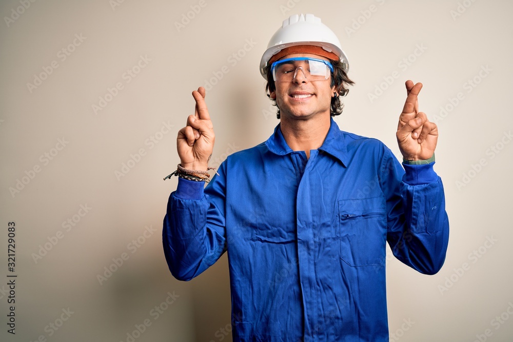 Young constructor man wearing uniform and security helmet over isolated white background gesturing finger crossed smiling with hope and eyes closed. Luck and superstitious concept. Young constructor man wearing uniform and security helmet over isolated white background gesturing finger crossed smiling with hope and eyes closed. Luck and superstitious concept.