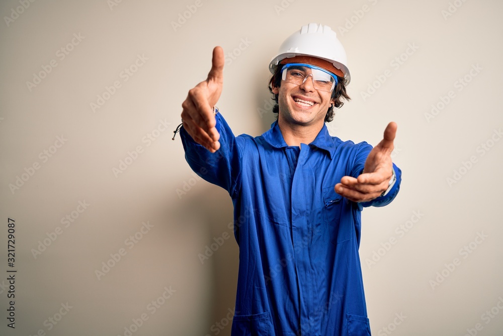 Young constructor man wearing uniform and security helmet over isolated white background looking at the camera smiling with open arms for hug. Cheerful expression embracing happiness. Young constructor man wearing uniform and security helmet over isolated white background looking at the camera smiling with open arms for hug. Cheerful expression embracing happiness.