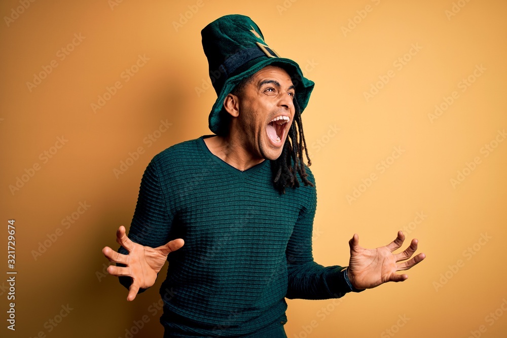 Young african american man wearing green hat with clover celebrating saint patricks day crazy and mad shouting and yelling with aggressive expression and arms raised. Frustration concept. Young african american man wearing green hat with clover celebrating saint patricks day crazy and mad shouting and yelling with aggressive expression and arms raised. Frustration concept.