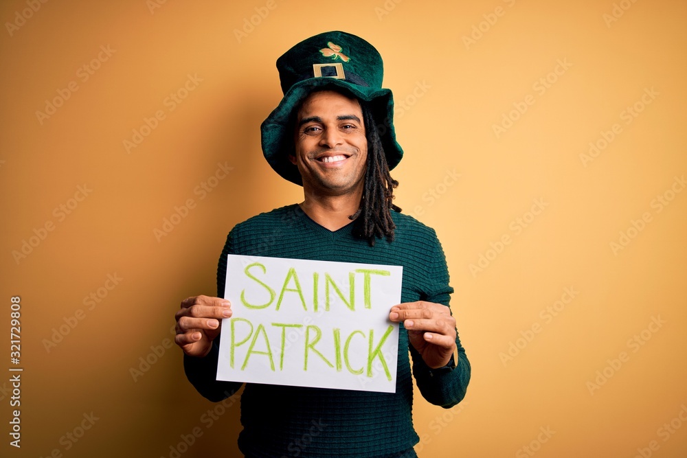 Young african american man wearing green hat holding banner celebrating saint patricks day with a happy face standing and smiling with a confident smile showing teeth Young african american man wearing green hat holding banner celebrating saint patricks day with a happy face standing and smiling with a confident smile showing teeth