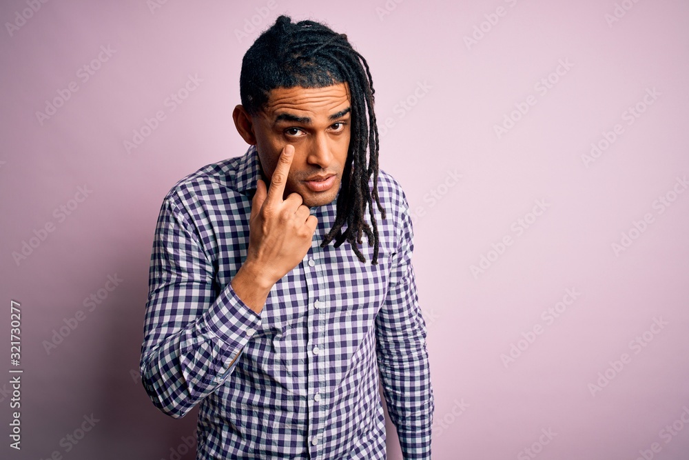 Young handsome african american afro man with dreadlocks wearing casual shirt Pointing to the eye watching you gesture, suspicious expression Young handsome african american afro man with dreadlocks wearing casual shirt Pointing to the eye watching you gesture, suspicious expression
