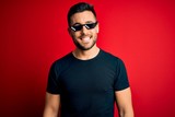 Young handsome man wearing funny thug life sunglasses over isolated red background with a happy and cool smile on face. Lucky person.
