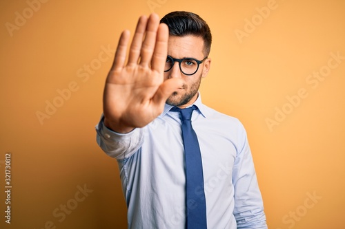Young handsome businessman wearing tie and glasses standing over yellow background doing stop sing with palm of the hand. Warning expression with negative and serious gesture on the face.